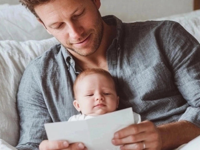  Father sitting on a bed with his baby in his lap, reading a card together, sharing a quiet and loving Father’s Day moment in a cosy setting.