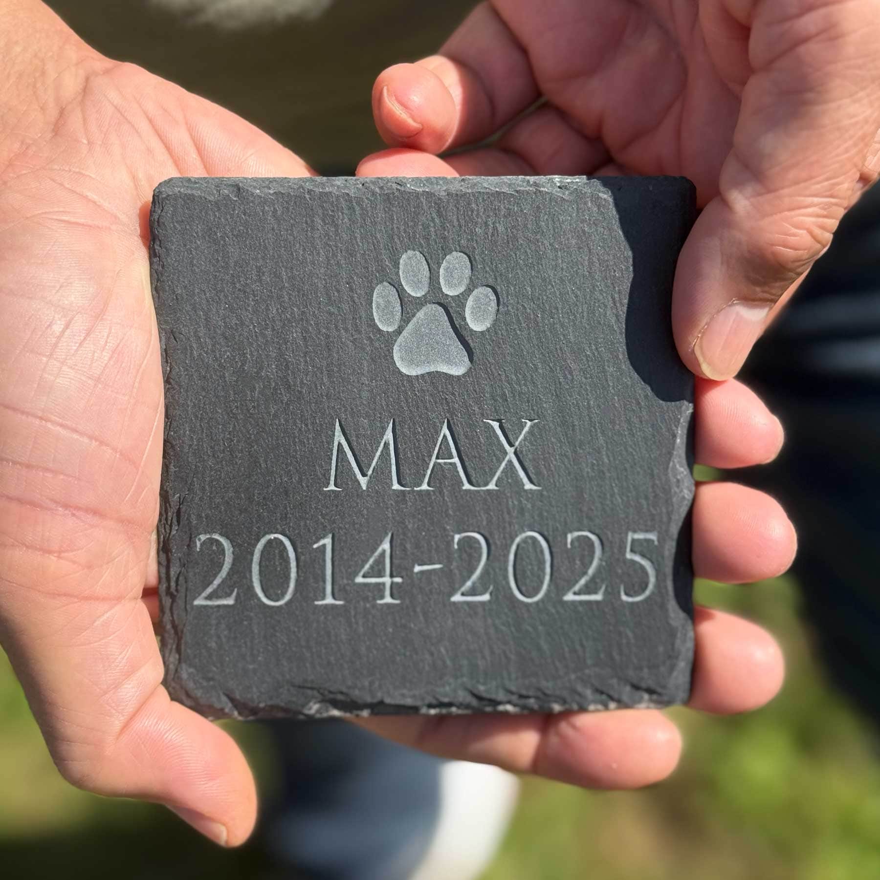 Person holding a slate stone with engraved paw print and text, set against a natural background.